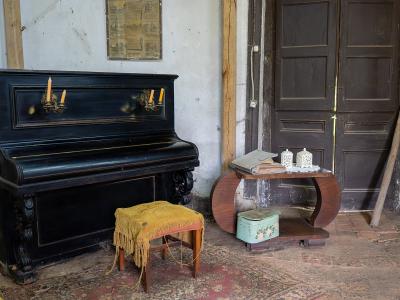 An interior room featuring a black upright piano with candle sconces, a yellow-fringed stool, a curved wooden side table with a vintage suitcase, and a portrait on the wall.