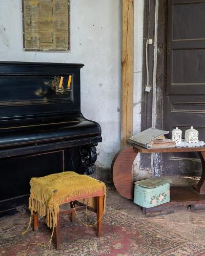 An interior room featuring a black upright piano with candle sconces, a yellow-fringed stool, a curved wooden side table with a vintage suitcase, and a portrait on the wall.