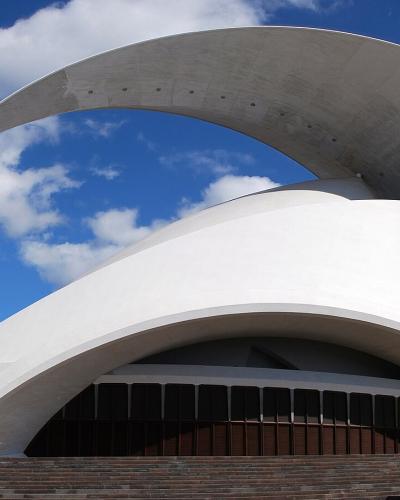 A low-angle view of the Auditorio de Tenerife, a modern white concert hall with a distinctive large curved roof structure, set against a blue sky with clouds.