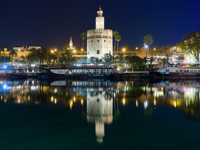 A night photograph of a large, illuminated twelve-sided stone tower situated on the bank of a river, with tour boats docked in the foreground and palm trees visible along the waterfront.