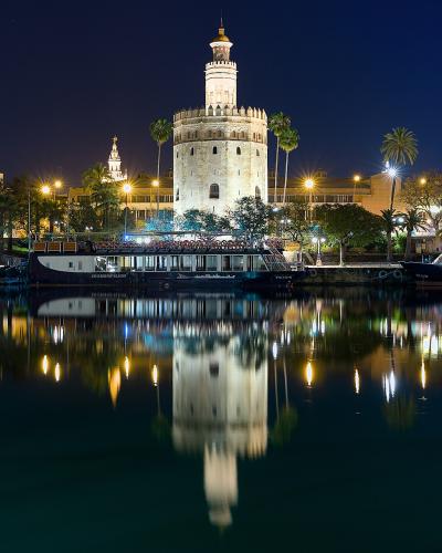 A night photograph of a large, illuminated twelve-sided stone tower situated on the bank of a river, with tour boats docked in the foreground and palm trees visible along the waterfront.