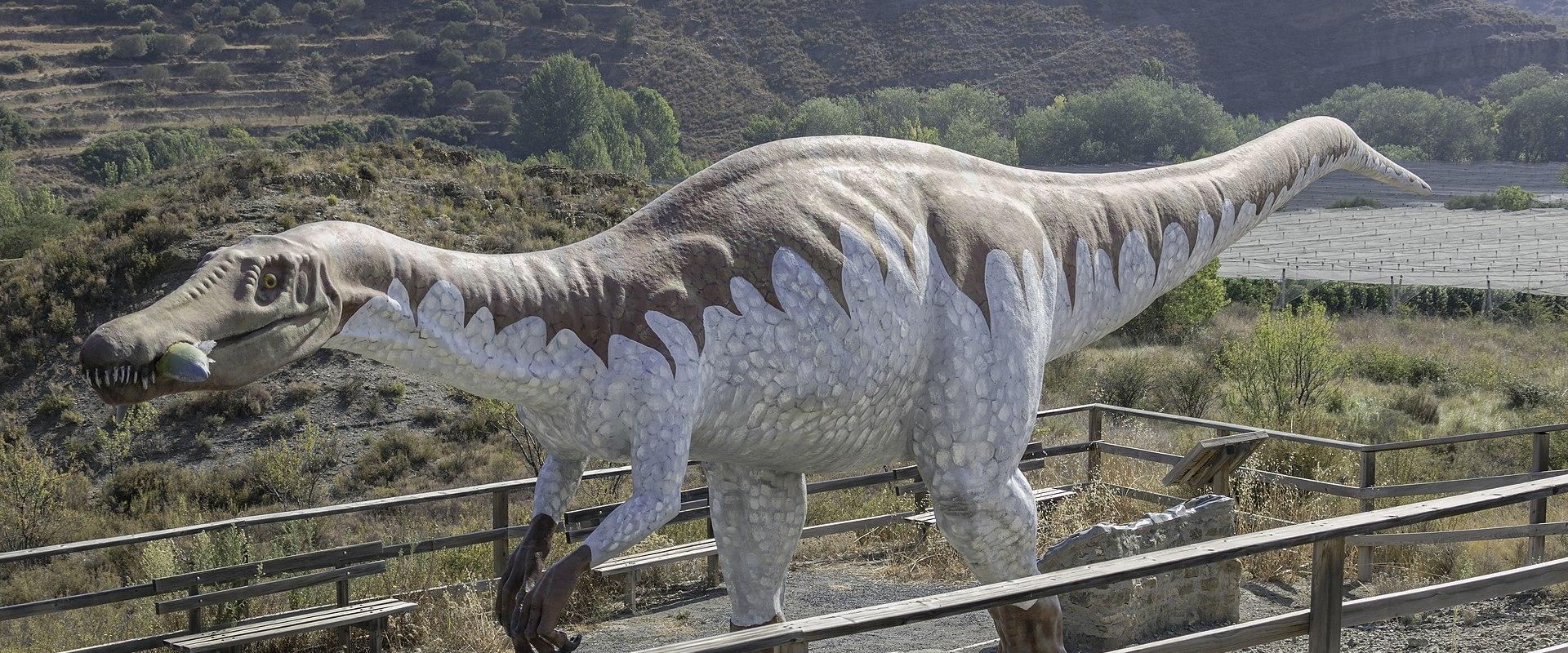 A large, life-sized model of a Baryonyx walkeri dinosaur statue stands on a gravel platform surrounded by wooden railings, with dry, hilly terrain and vineyards in the background.