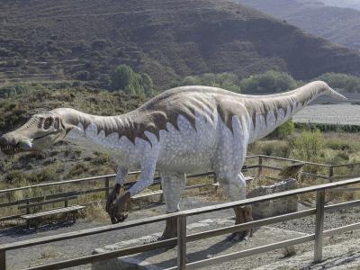 A large, life-sized model of a Baryonyx walkeri dinosaur statue stands on a gravel platform surrounded by wooden railings, with dry, hilly terrain and vineyards in the background.