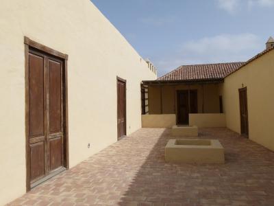 A sunlit interior courtyard of a traditional Canarian-style building with stucco walls, wooden doors, terracotta tile flooring, and two rectangular stone planters or water basins in the center.