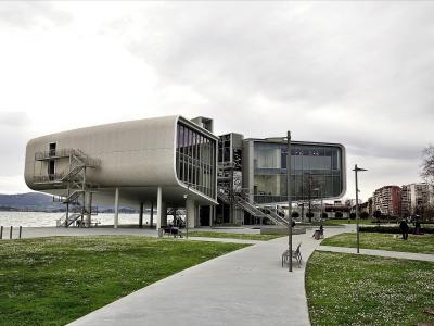 A modern, white, box-like building with a distinctive cantilevered section and external metal staircases, situated on a waterfront promenade next to a body of water with hills in the background.