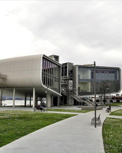 A modern, white, box-like building with a distinctive cantilevered section and external metal staircases, situated on a waterfront promenade next to a body of water with hills in the background.
