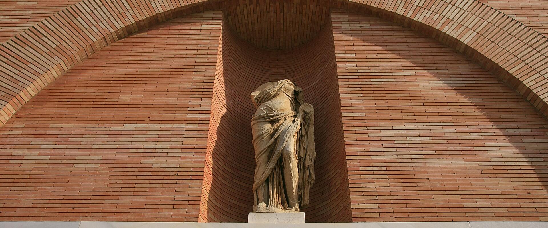 A close-up of a brick facade featuring a large archway with a classical statue inside, and the word "MVSEO" in large gold letters on a stone ledge below.