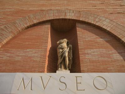 A close-up of a brick facade featuring a large archway with a classical statue inside, and the word "MVSEO" in large gold letters on a stone ledge below.