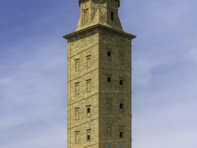 A tall, ancient Roman lighthouse stands atop a grassy promontory overlooking a rocky coastline and the ocean, with a town visible in the distance to the left.