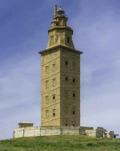 A tall, ancient Roman lighthouse stands atop a grassy promontory overlooking a rocky coastline and the ocean, with a town visible in the distance to the left.
