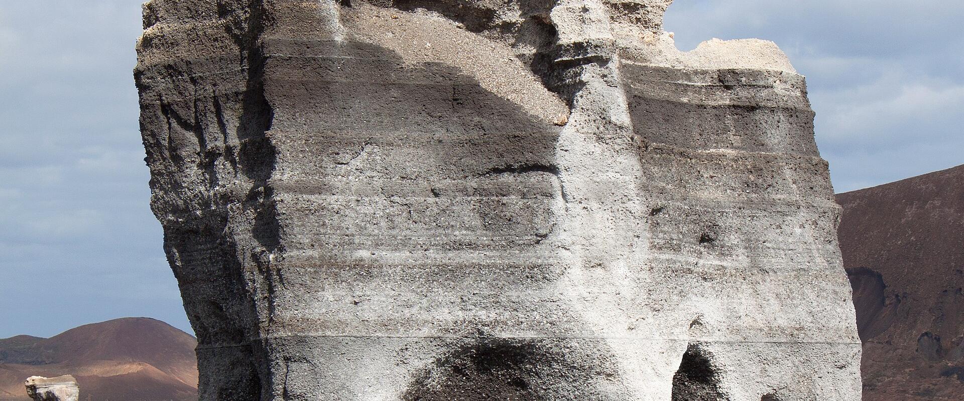 A large, eroded volcanic rock formation with distinct horizontal layers of dark tephra and lighter ash, set in a barren landscape with other volcanic cones in the background.