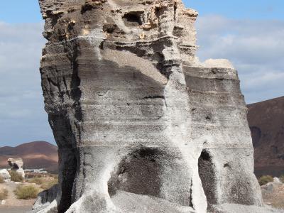 A large, eroded volcanic rock formation with distinct horizontal layers of dark tephra and lighter ash, set in a barren landscape with other volcanic cones in the background.