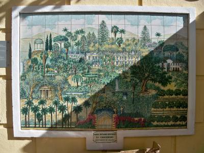 A large ceramic tile mural depicting a landscape with palm trees, a mansion, and a temple, mounted on a yellow wall with a small plaque below it.