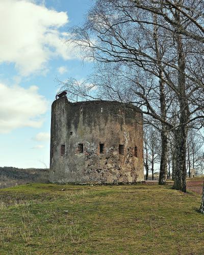 A weathered, cylindrical stone tower with narrow vertical openings stands on a grassy knoll, framed by bare deciduous trees under a partly cloudy sky.