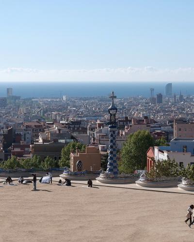 A wide-angle view of a large, open plaza with distinctive mosaic-tiled benches and a tall, blue-and-white checkered lamp post, overlooking a dense cityscape and the sea in the distance.