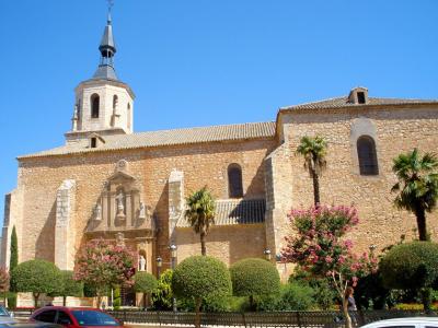 A stone church with a bell tower and spire, featuring a sculpted facade with statues, surrounded by manicured trees and flowering bushes under a clear blue sky.