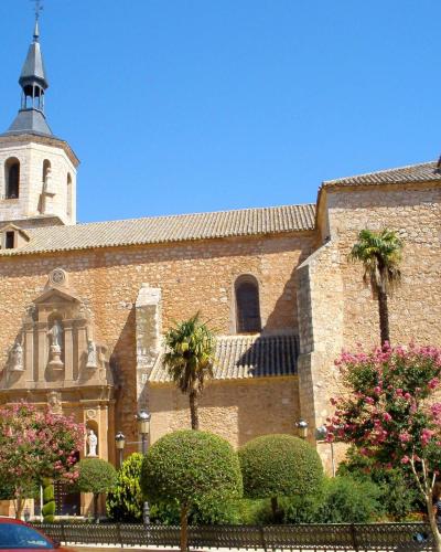 A stone church with a bell tower and spire, featuring a sculpted facade with statues, surrounded by manicured trees and flowering bushes under a clear blue sky.