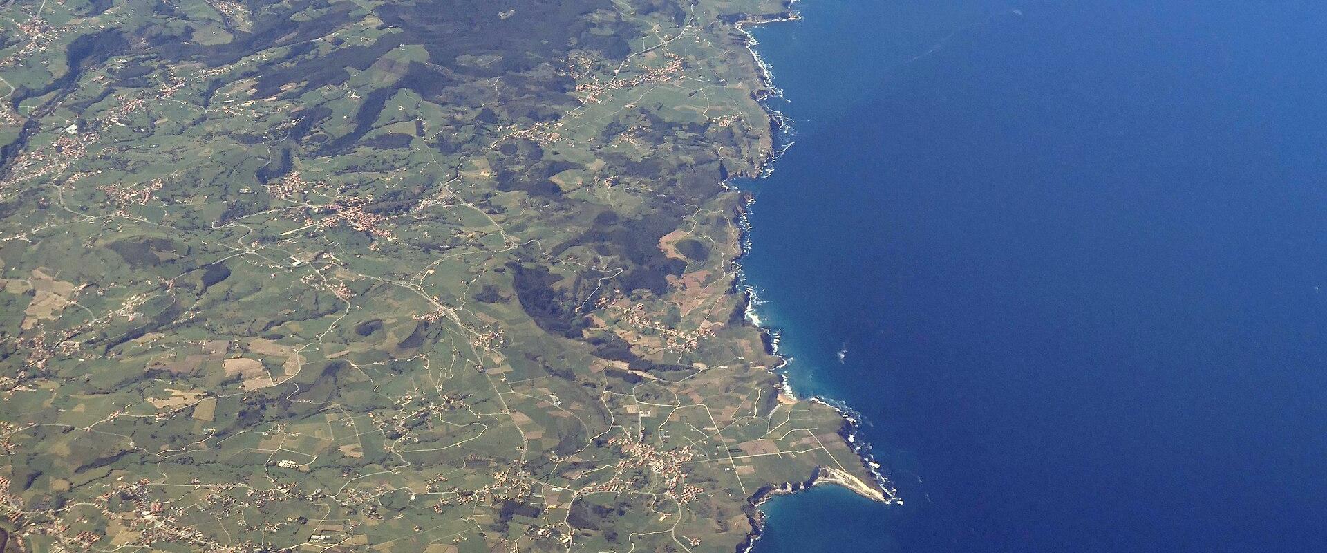 An aerial photograph showing a green, hilly coastline with a river estuary and coastal towns meeting the blue sea.