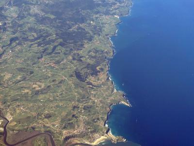 An aerial photograph showing a green, hilly coastline with a river estuary and coastal towns meeting the blue sea.