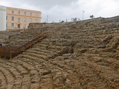 The image shows the excavated stone seating tiers of an ancient Roman theatre ruin, with modern wooden walkways and stairs installed for visitors, and a large pale yellow building in the background.