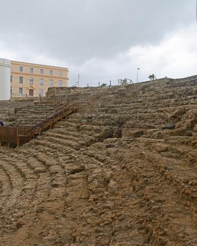 The image shows the excavated stone seating tiers of an ancient Roman theatre ruin, with modern wooden walkways and stairs installed for visitors, and a large pale yellow building in the background.