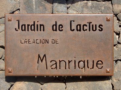 A rusty metal sign mounted on a volcanic rock wall, reading "Jardín de Cactus" and "CREACION DE Manrique".