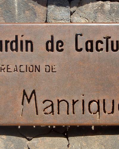 A rusty metal sign mounted on a volcanic rock wall, reading "Jardín de Cactus" and "CREACION DE Manrique".