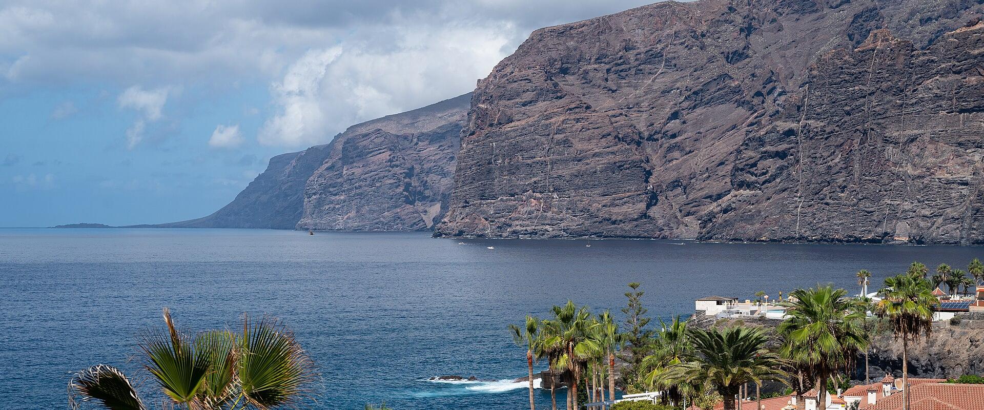 A view of massive, dark volcanic sea cliffs rising directly from the ocean, with a foreground of residential rooftops and palm trees.