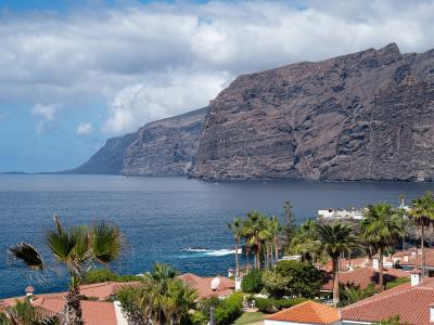A view of massive, dark volcanic sea cliffs rising directly from the ocean, with a foreground of residential rooftops and palm trees.