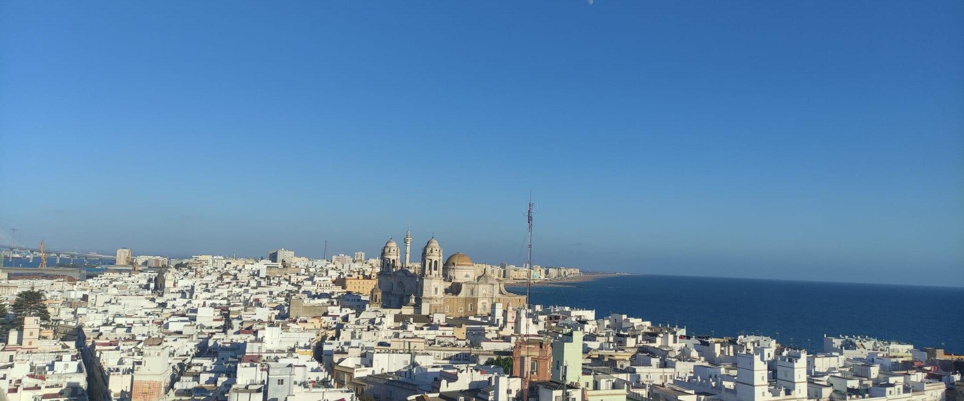 A panoramic daytime view of the white-washed city of Cádiz, featuring the Cádiz Cathedral prominently in the center, with the Atlantic Ocean to the right and the Bay of Cádiz to the left.