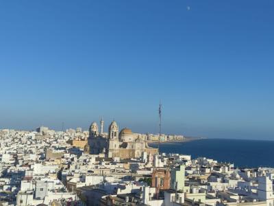 A panoramic daytime view of the white-washed city of Cádiz, featuring the Cádiz Cathedral prominently in the center, with the Atlantic Ocean to the right and the Bay of Cádiz to the left.