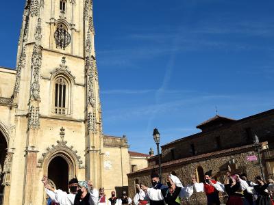 A group of people in traditional folk costumes dancing in a paved plaza in front of a tall, ornate Gothic church tower under a clear blue sky.