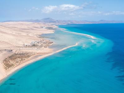 An aerial view of a long, curved sandy spit separating a shallow turquoise lagoon from the deep blue ocean, with arid dunes and distant mountains in the background.