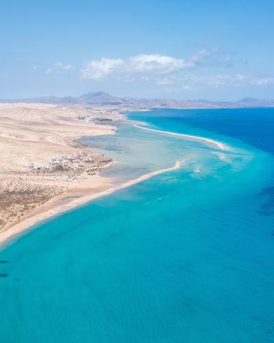 An aerial view of a long, curved sandy spit separating a shallow turquoise lagoon from the deep blue ocean, with arid dunes and distant mountains in the background.