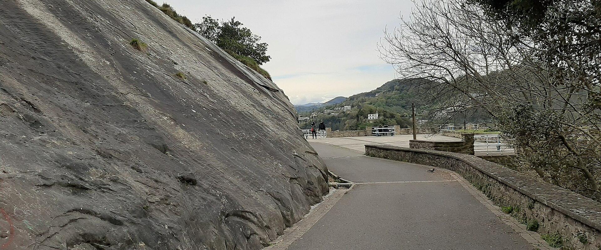 A paved walkway curving to the right, bordered on the left by a large, steep, grey rock face and on the right by a low stone wall, with a view of a bay and distant hills in the background.