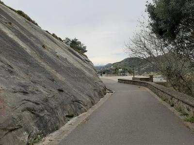 A paved walkway curving to the right, bordered on the left by a large, steep, grey rock face and on the right by a low stone wall, with a view of a bay and distant hills in the background.