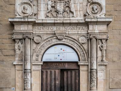 The image shows the ornate stone entrance facade of a building with a large arched doorway, flanked by columns and topped with sculptural reliefs and a coat of arms; the inscription "MUSEO DE NAVARRA" is clearly carved above the arch, and a modern po