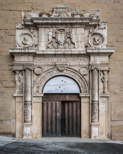 The image shows the ornate stone entrance facade of a building with a large arched doorway, flanked by columns and topped with sculptural reliefs and a coat of arms; the inscription "MUSEO DE NAVARRA" is clearly carved above the arch, and a modern po