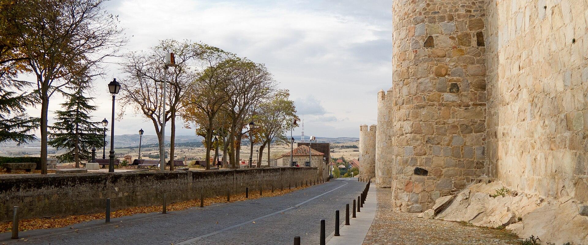 A cobblestone walkway runs alongside a high medieval stone wall with square towers and crenellations, bordered by trees and a low wall overlooking a valley.