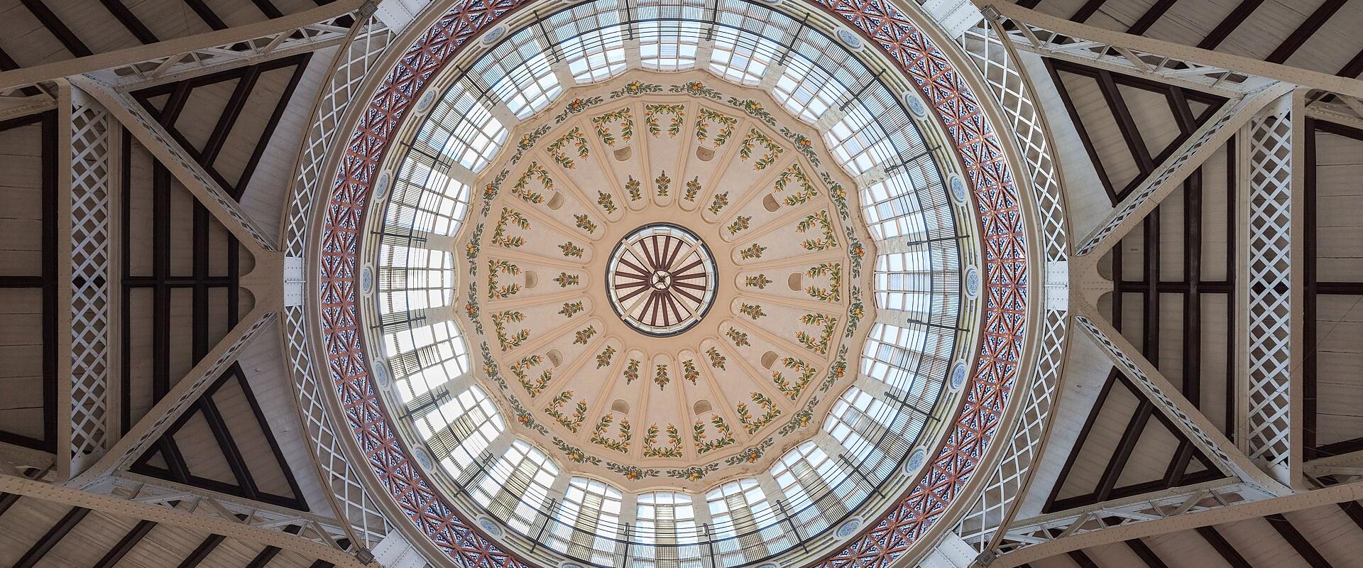 An upward view of an ornate, circular glass dome ceiling with decorative painted panels and a surrounding iron-and-glass structural framework.