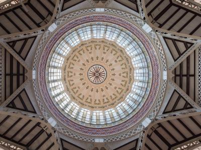 An upward view of an ornate, circular glass dome ceiling with decorative painted panels and a surrounding iron-and-glass structural framework.