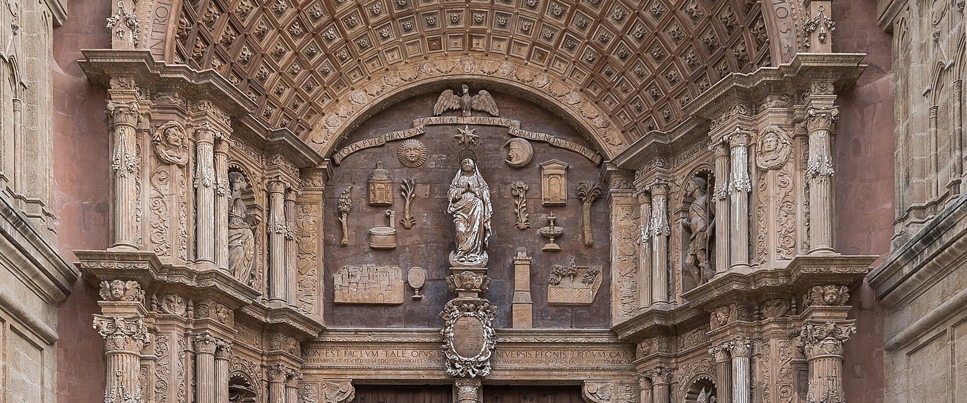 A close-up view of the ornate, Renaissance-style main entrance (Portal de la Portalada) of a large Gothic cathedral, featuring a massive wooden door, a coffered archway with Latin inscriptions, and a central statue of the Virgin Mary.