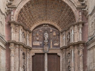 A close-up view of the ornate, Renaissance-style main entrance (Portal de la Portalada) of a large Gothic cathedral, featuring a massive wooden door, a coffered archway with Latin inscriptions, and a central statue of the Virgin Mary.