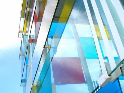 A low-angle architectural detail of a modern building featuring a facade of colorful, translucent glass panels (blue, yellow, red) supported by white structural beams against a blue sky.