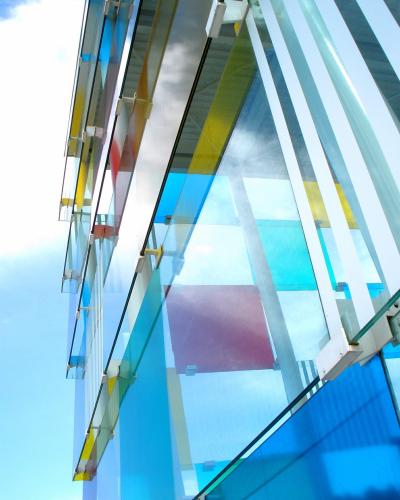 A low-angle architectural detail of a modern building featuring a facade of colorful, translucent glass panels (blue, yellow, red) supported by white structural beams against a blue sky.