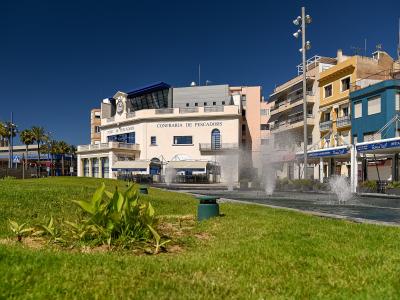 A waterfront plaza with a grassy foreground, fountains, and a prominent building labeled "CONFRARIA DE PESCADORS" and "PORT DE PESCADORS" next to a restaurant area.