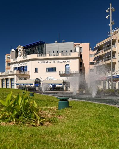 A waterfront plaza with a grassy foreground, fountains, and a prominent building labeled "CONFRARIA DE PESCADORS" and "PORT DE PESCADORS" next to a restaurant area.