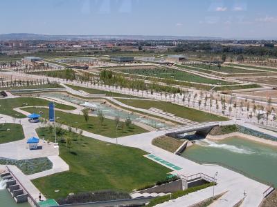 An aerial view of a large, modern landscaped park featuring winding water channels, concrete walkways, grassy areas, and distant urban development under a clear sky.