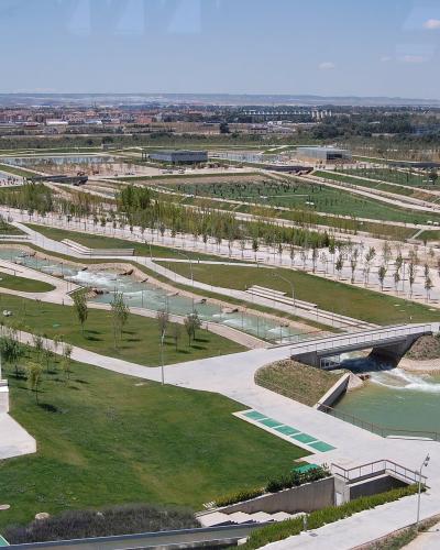 An aerial view of a large, modern landscaped park featuring winding water channels, concrete walkways, grassy areas, and distant urban development under a clear sky.