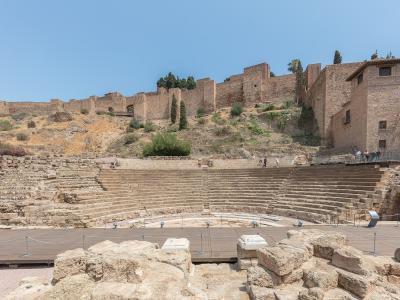 The image shows the stone seating tiers of an ancient Roman theatre with a modern wooden stage platform in the foreground, set against a hillside topped by the Alcazaba fortress walls.